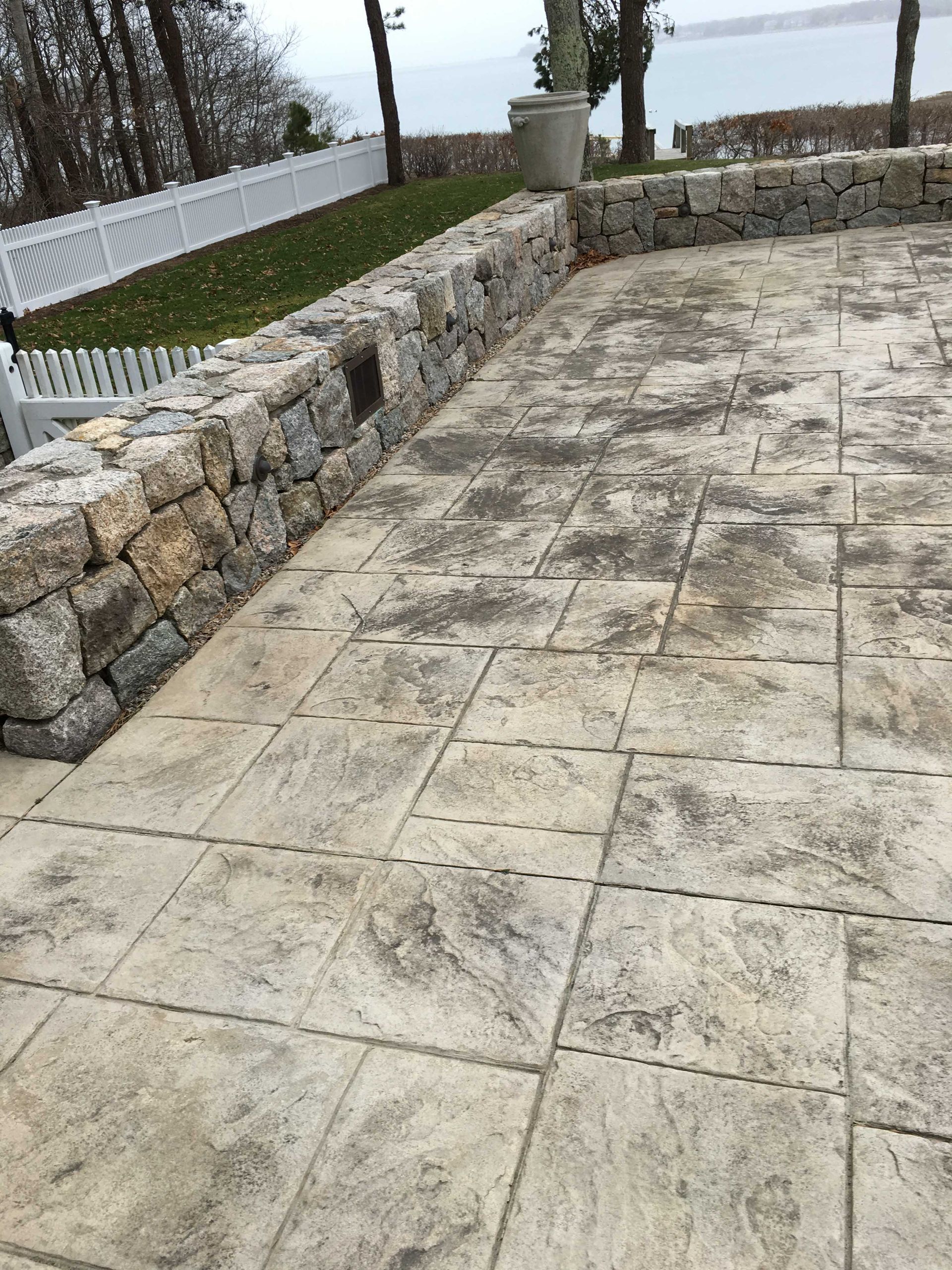 A stone patio beside a low stacked-stone retaining wall, with a white fence and water view in the background.