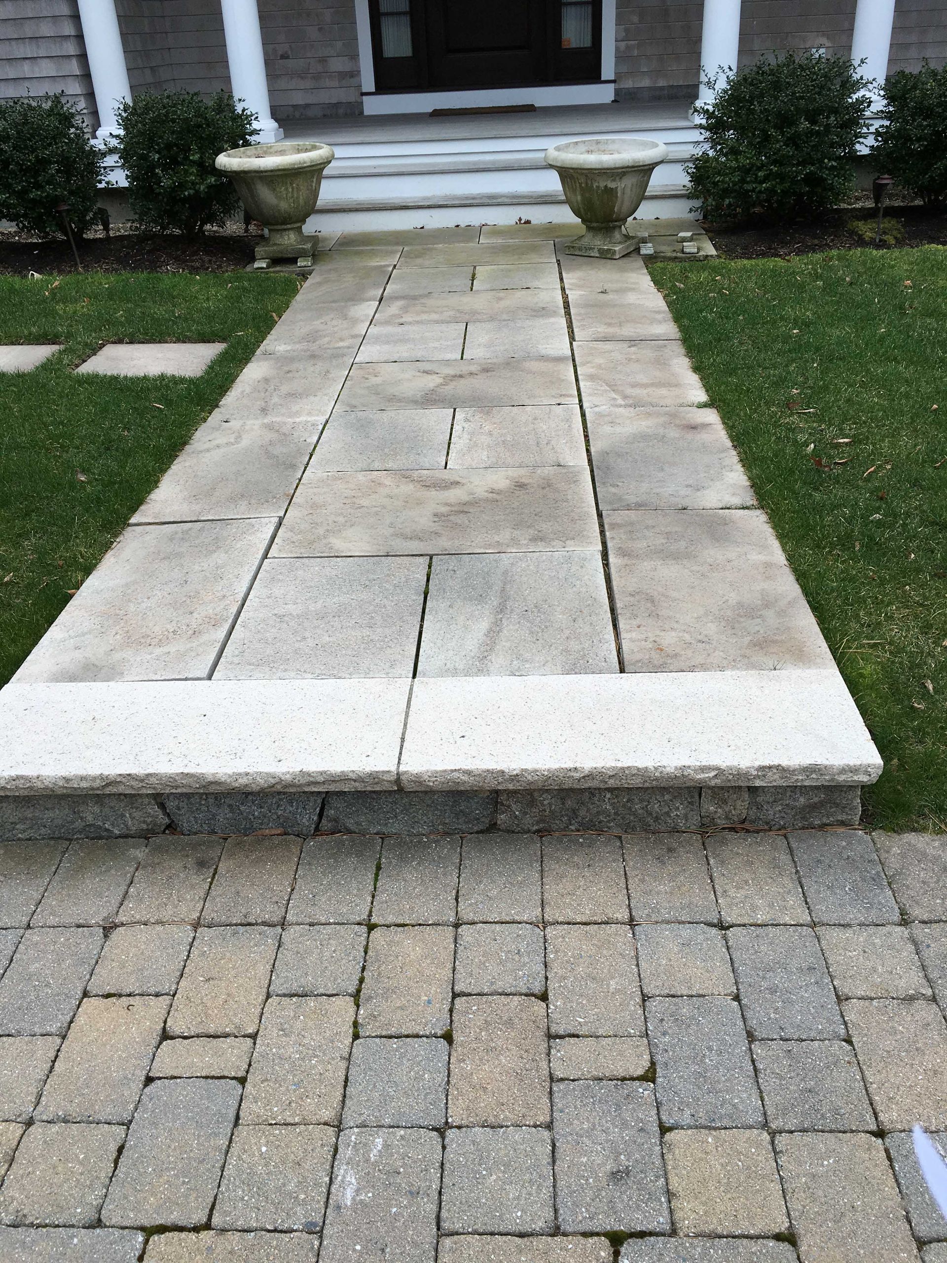 Stone walkway leading to a home entrance with two stone planters, grass, and a paved brick patio in the foreground.