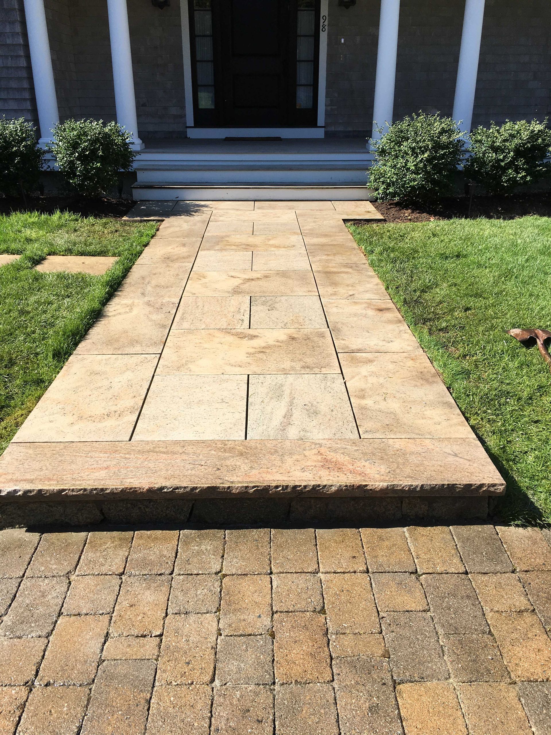 A paved walkway made of large tan stone slabs leads to the front steps and entrance of a residential home.
