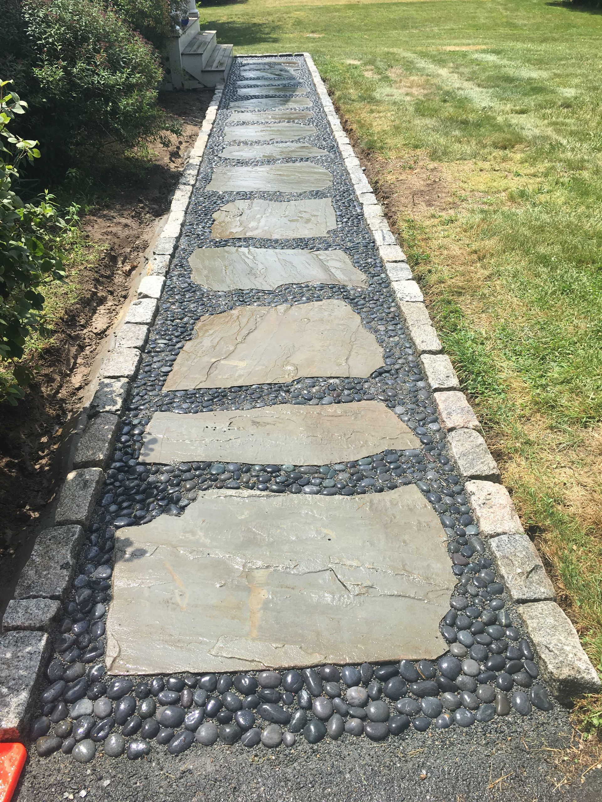 A garden path made of large, flat gray stepping stones framed by dark, round river stones and granite edges.