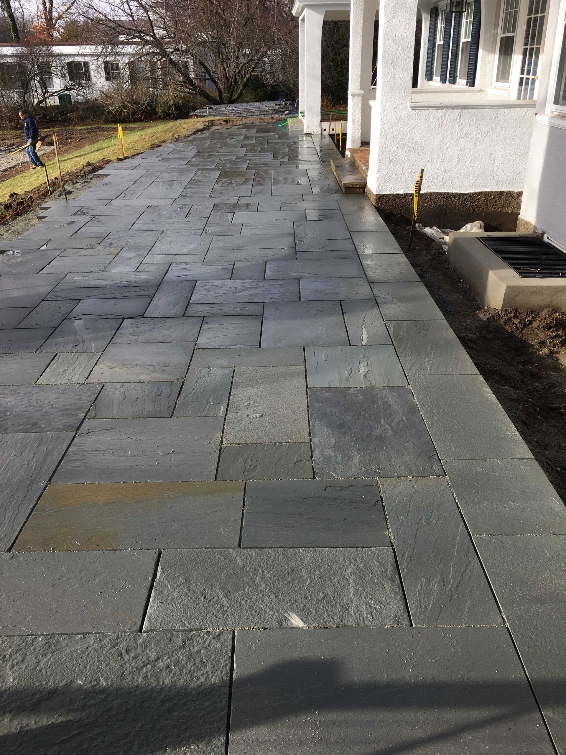 A paved walkway made of rectangular dark grey stone slabs leading along the side of a white house.