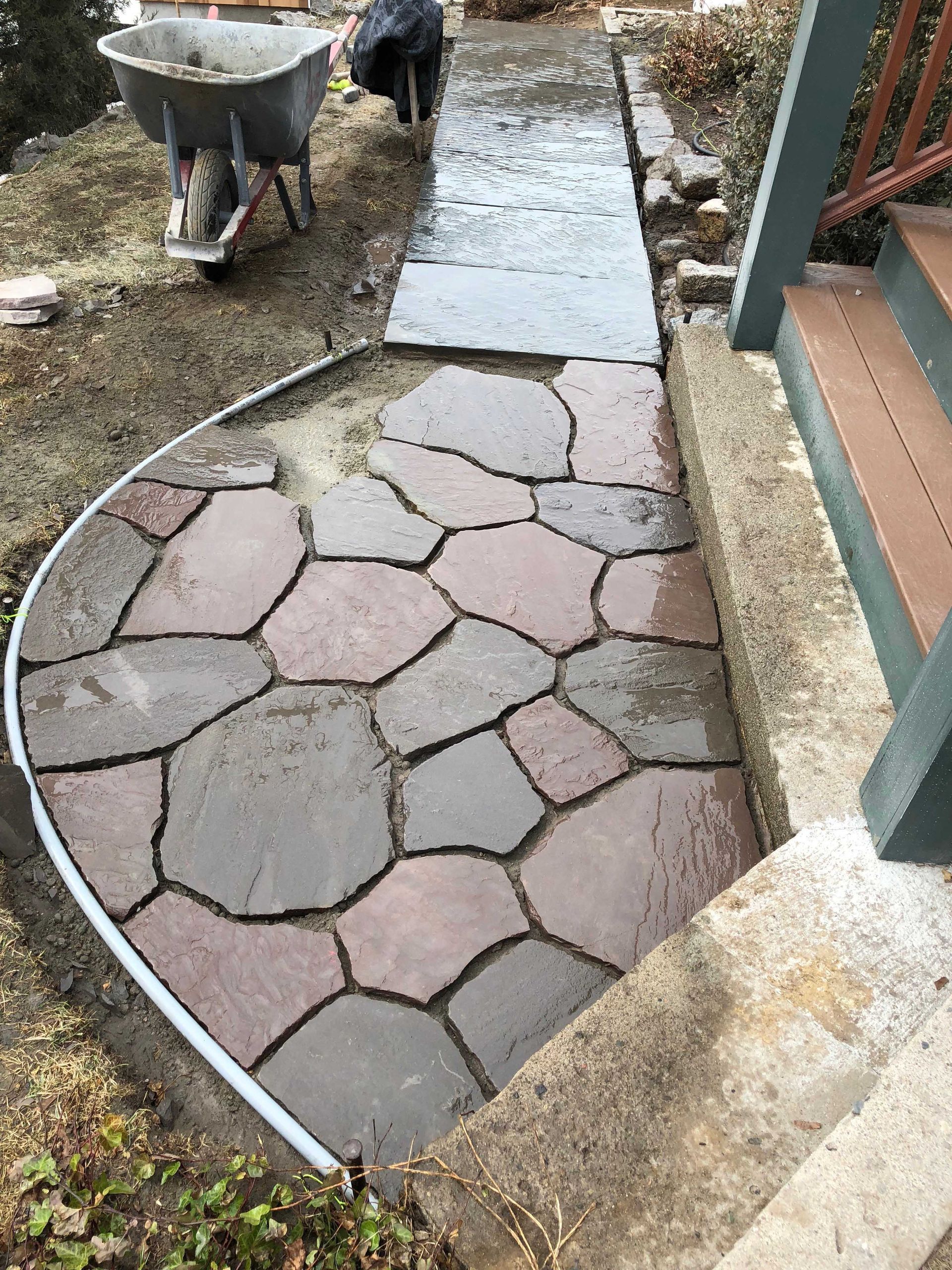 A stone patio being installed, with irregular red-brown flagstones arranged in a curve next to a wooden deck and wheelbarrow.
