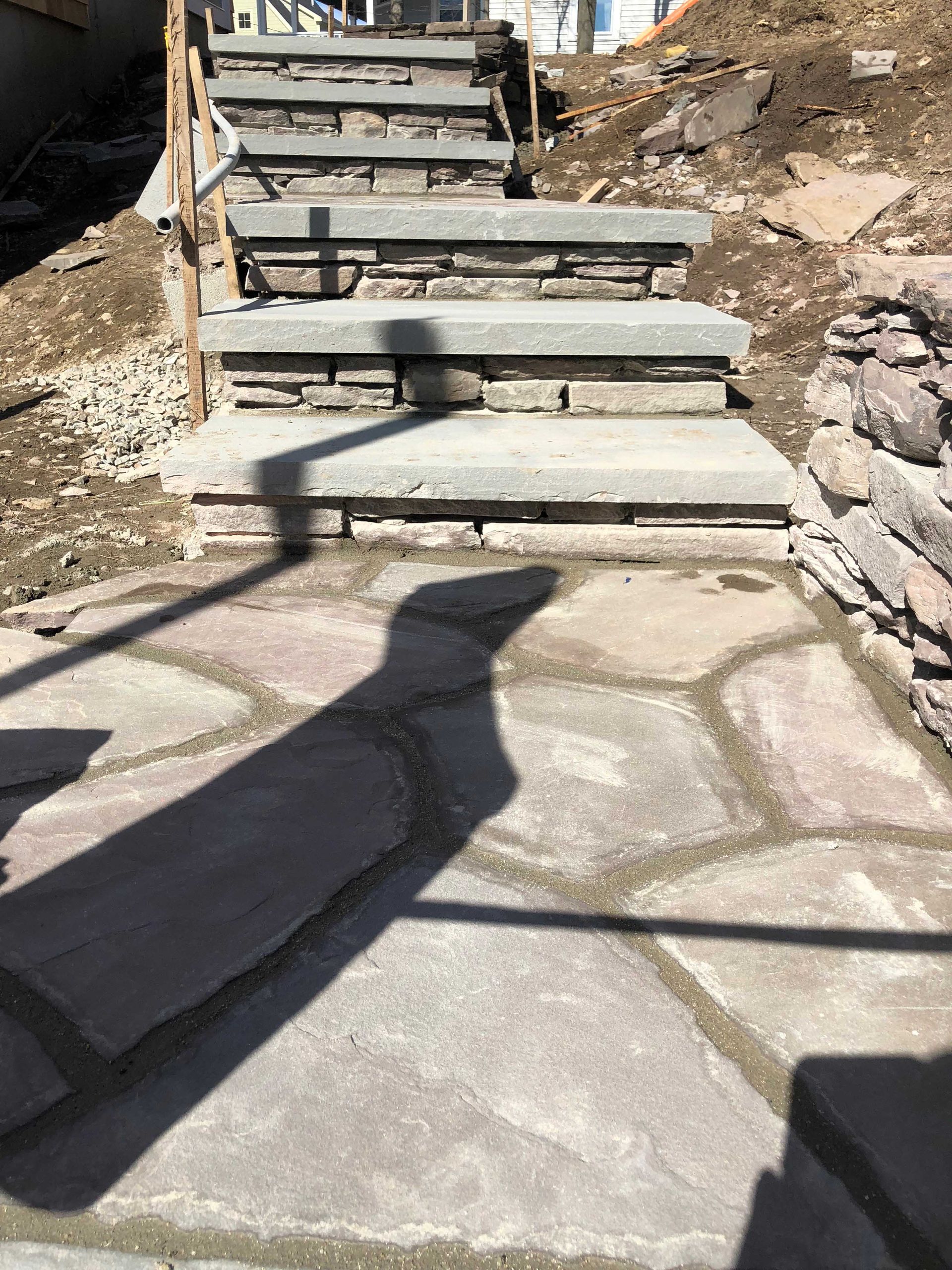 Stone stairs leading upward from a patio area, with a shadow cast across the path.