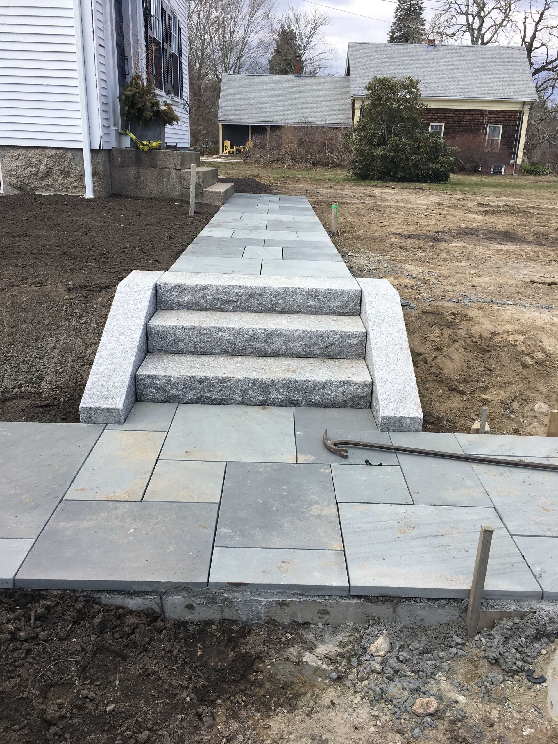 A new bluestone walkway with granite steps leads toward a house. The surrounding yard is under construction.