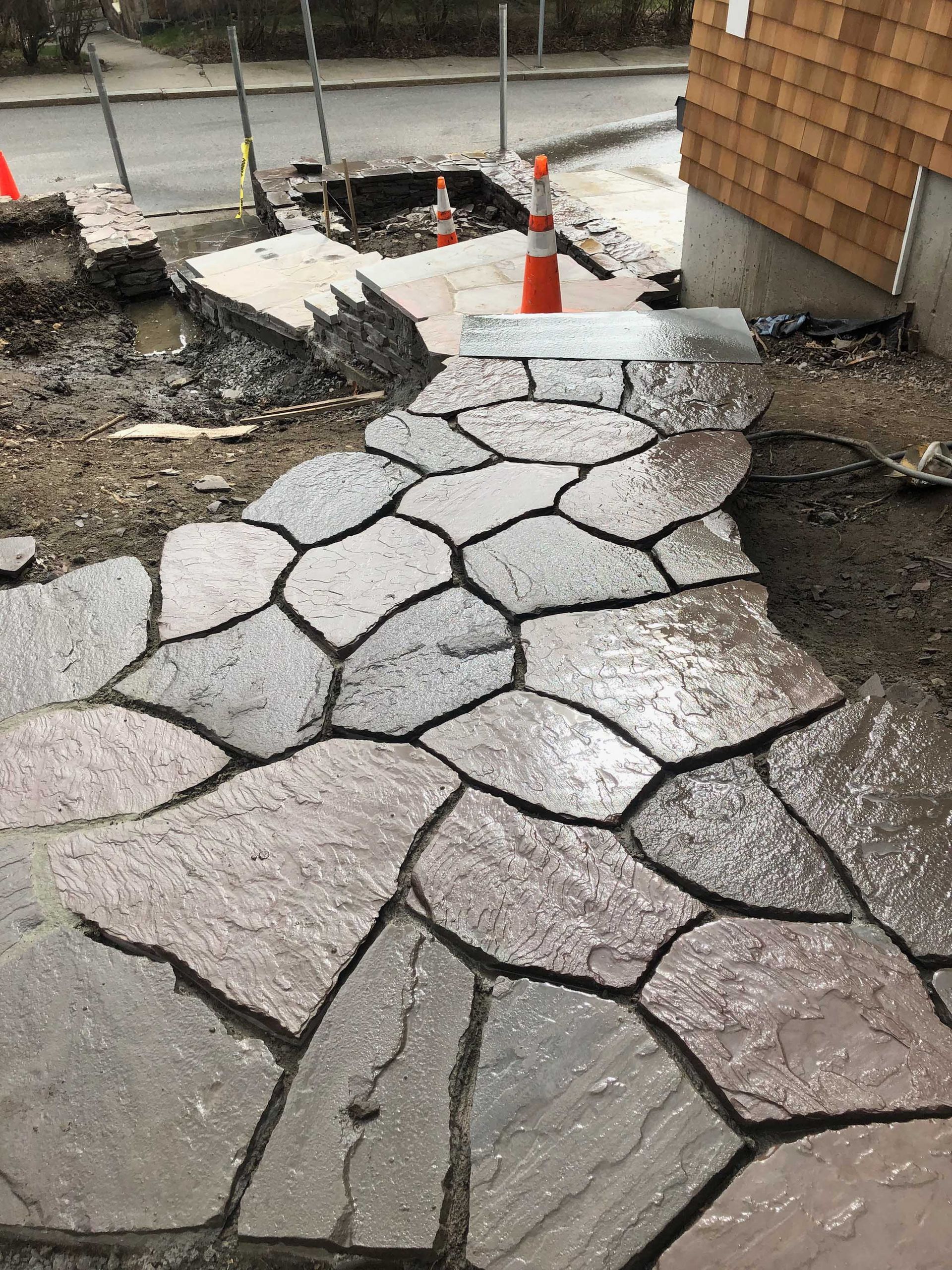 A newly laid stone path leads to a house foundation, with an orange traffic cone placed near a construction area.