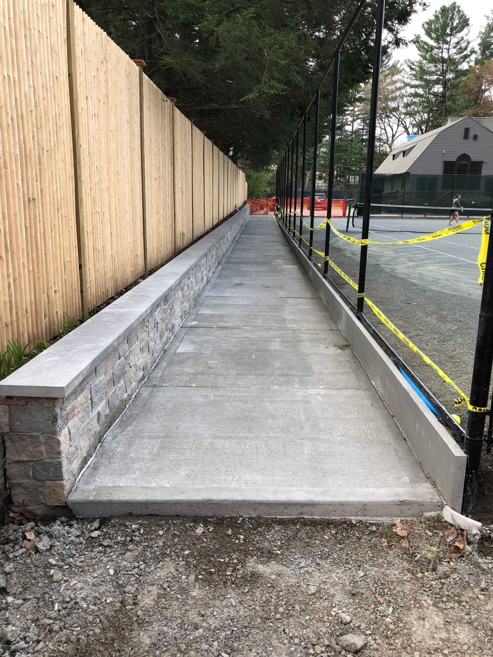 A sidewalk bordered by a stone wall and fence leads through a construction site with yellow caution tape.