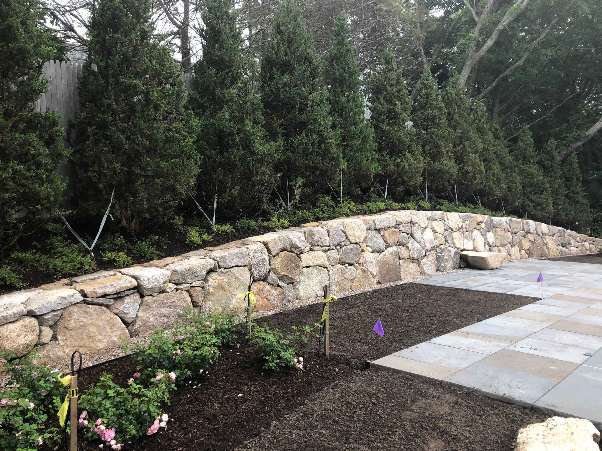 A stone retaining wall curves along a patio with a garden bed in the foreground and tall evergreen trees in the background.