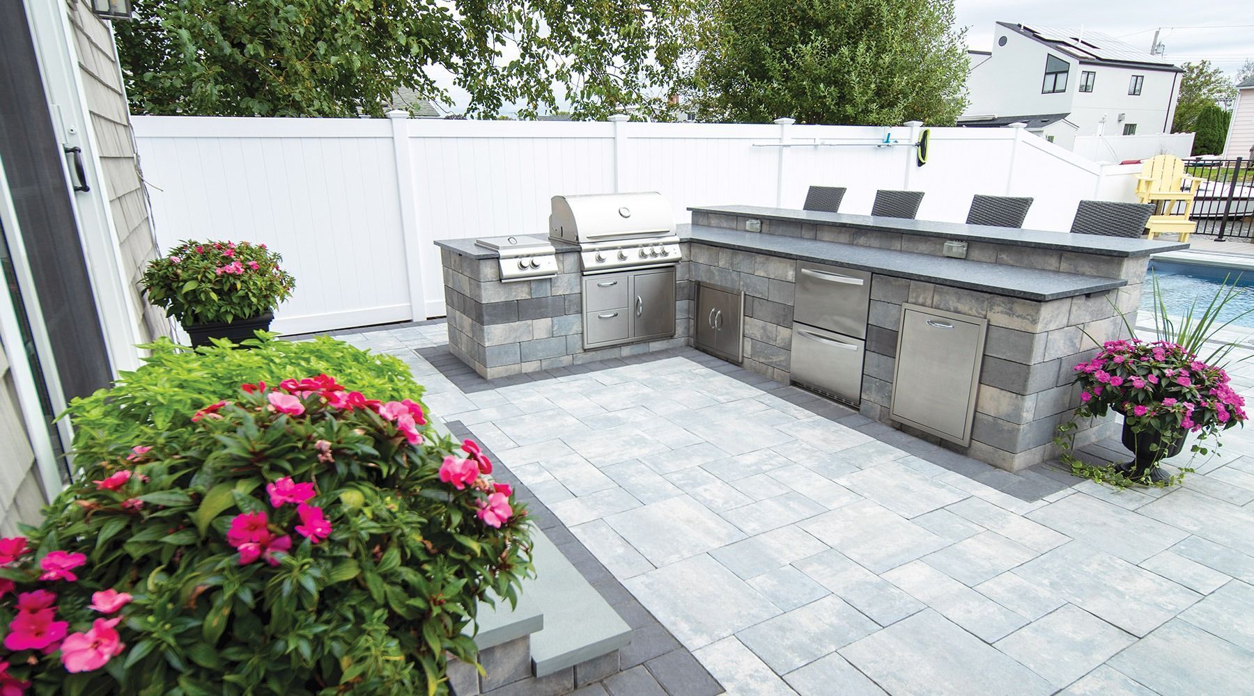 A patio with a grill and flowers in front of a pool.