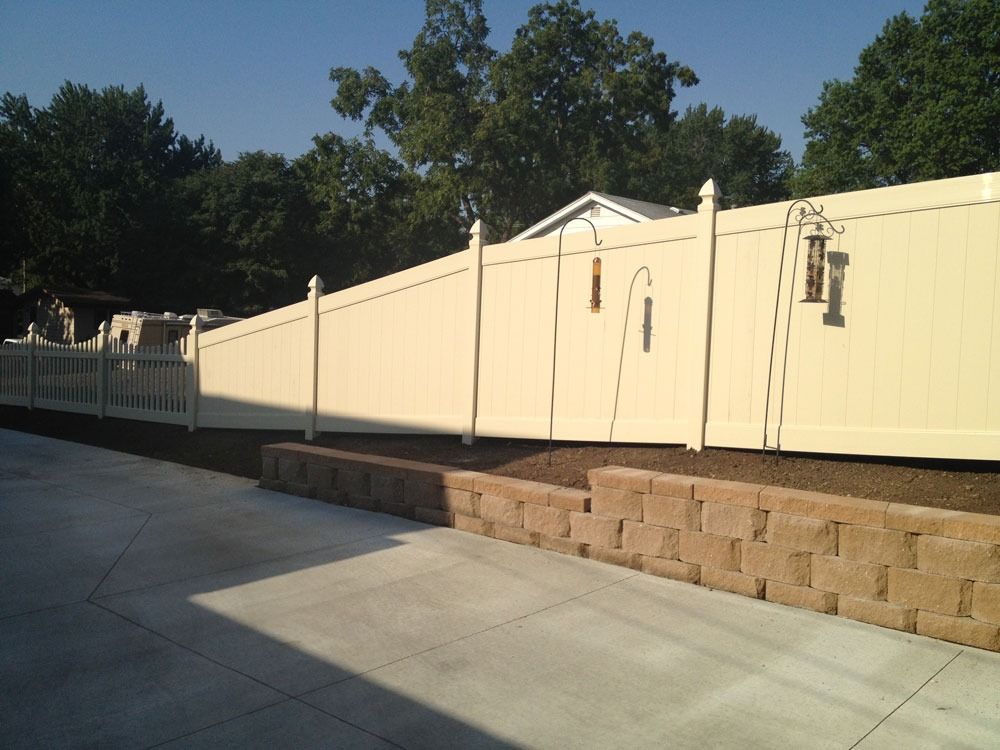 Beige vinyl fence along a concrete driveway with a retaining wall; trees in background.