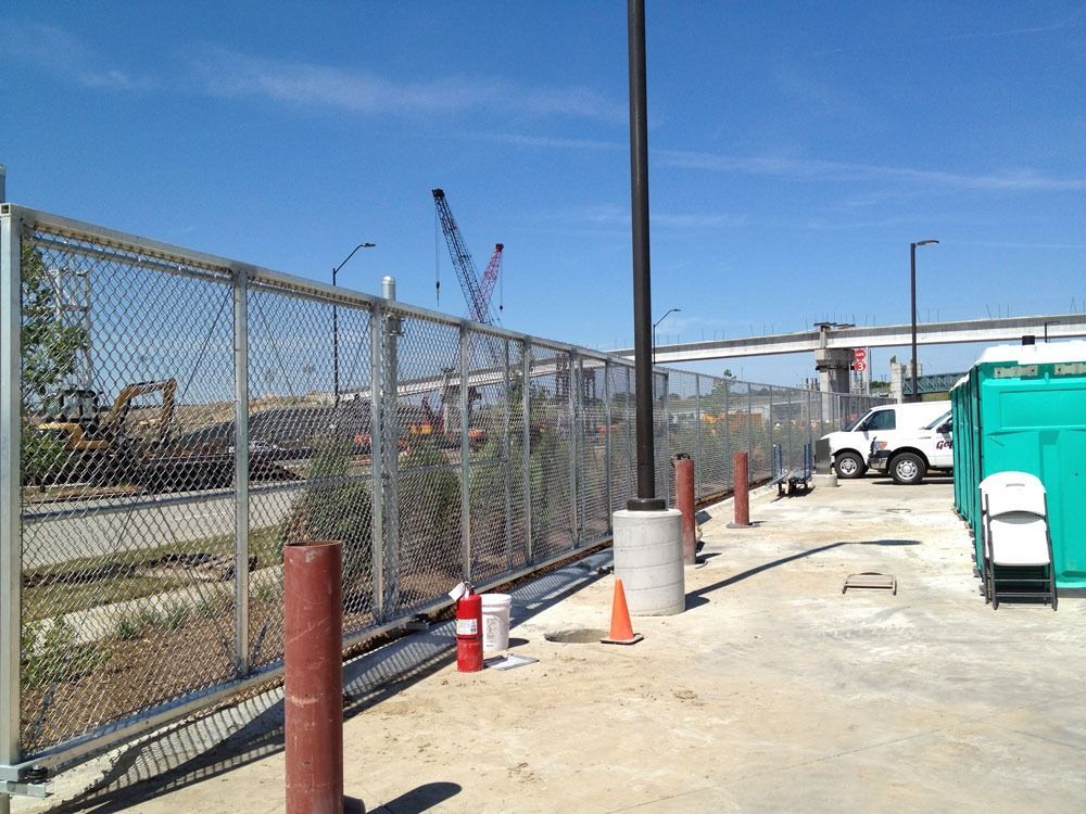 Chain-link fence at a construction site, with crane, trucks, and portable restrooms. Blue sky.