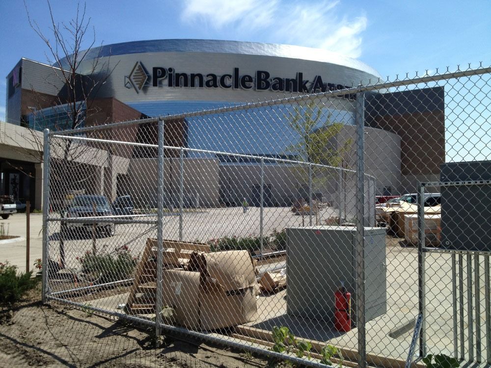 Chain-link fence in front of Pinnacle Bank Arena. Construction materials and an electrical box are visible. Blue sky in the background.