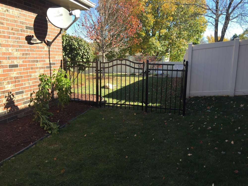 Black metal gate in a backyard, beside a brick wall and white fence, with green grass and fall foliage.