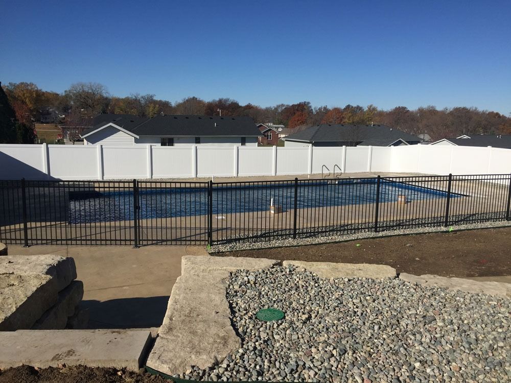 Swimming pool fenced with black bars, surrounded by white privacy fence, clear blue sky.