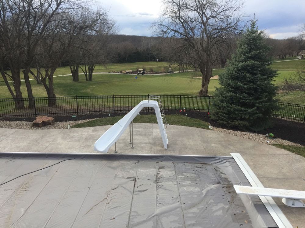 Backyard pool with a slide, overlooking a golf course. Pool cover on; trees in the background.