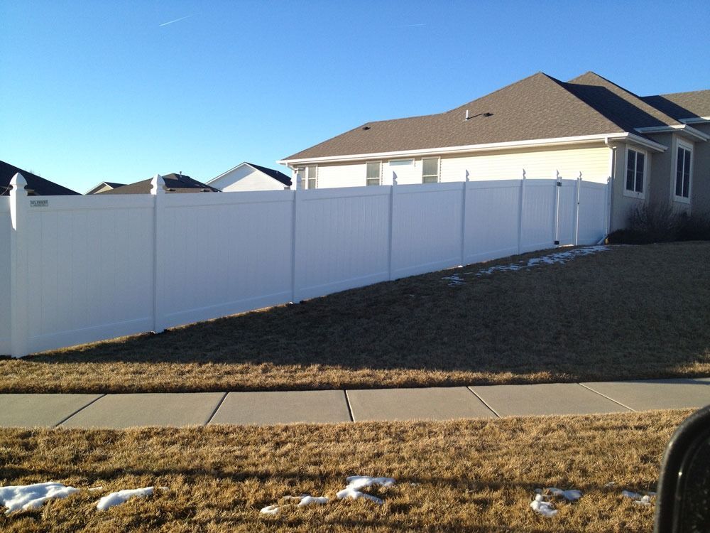 White vinyl fence around a house on a slight hill, sidewalk in foreground, blue sky.