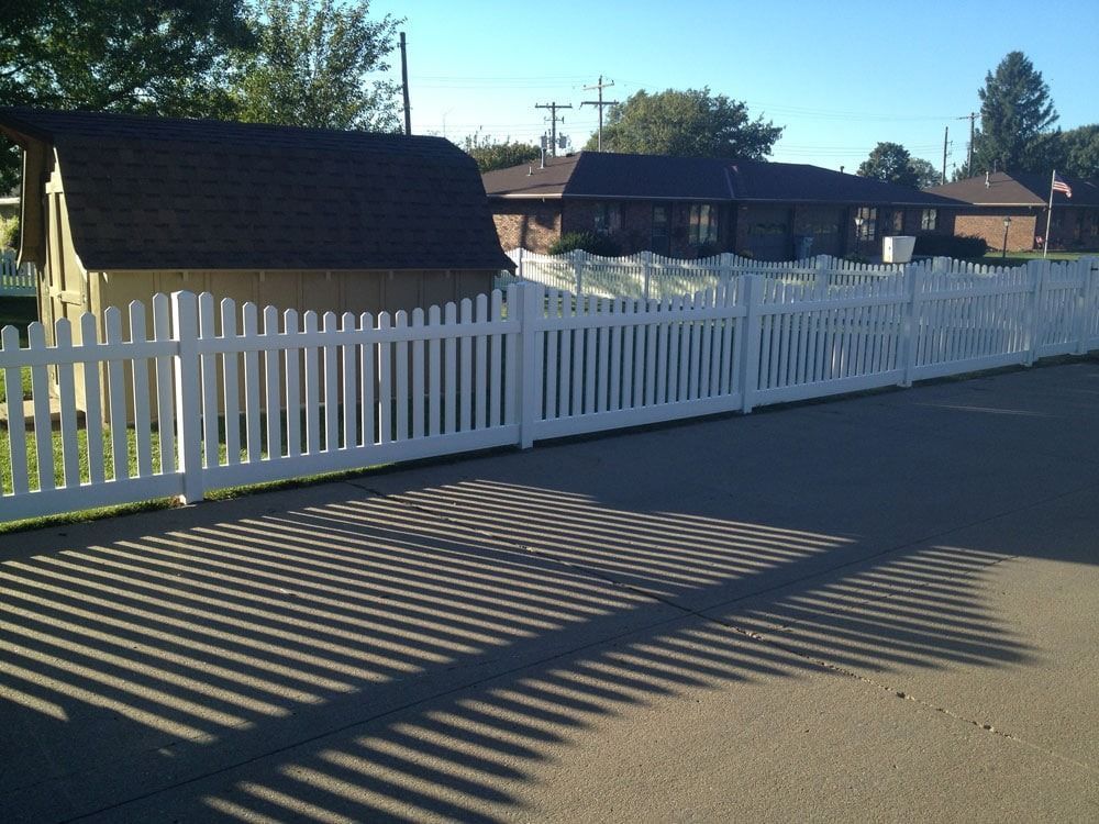 White picket fence in front of a shed and brick houses casting long shadows on a paved surface.