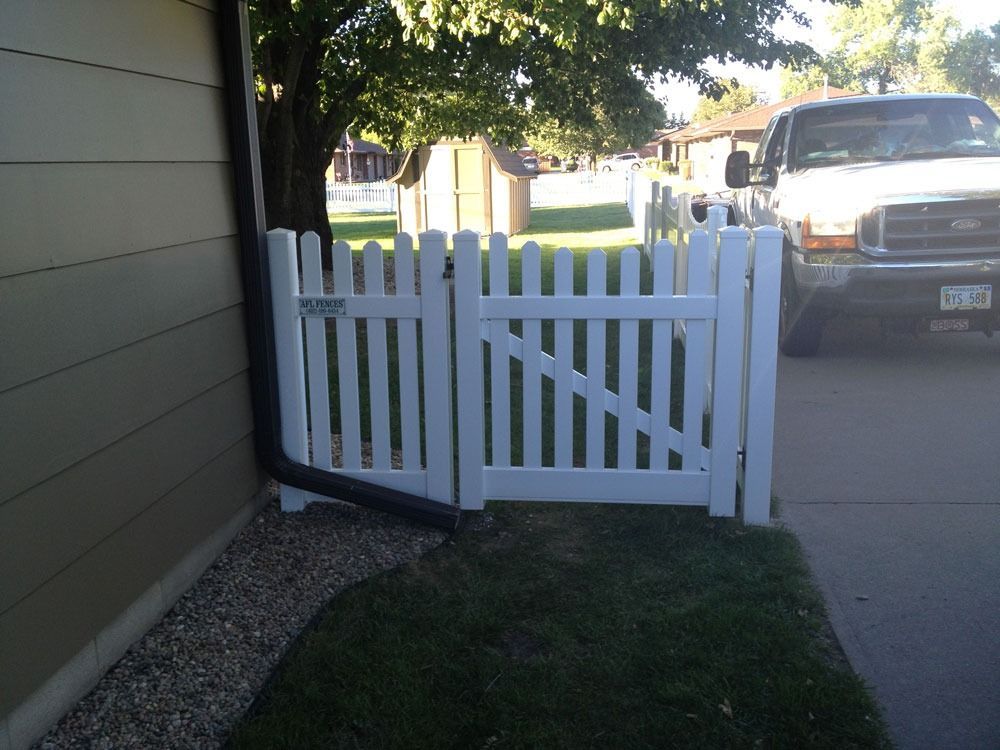 White picket fence and gate next to a house with a black downspout; a truck is parked nearby.