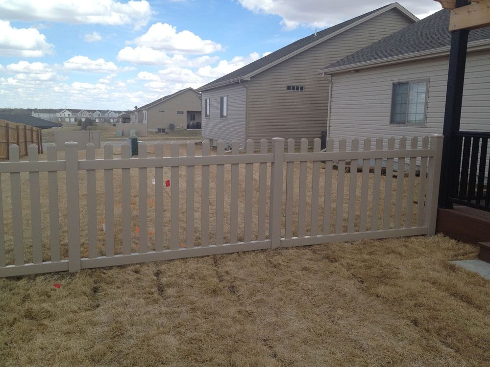 Tan picket fence in a yard with dry grass, with houses and blue sky in the background.