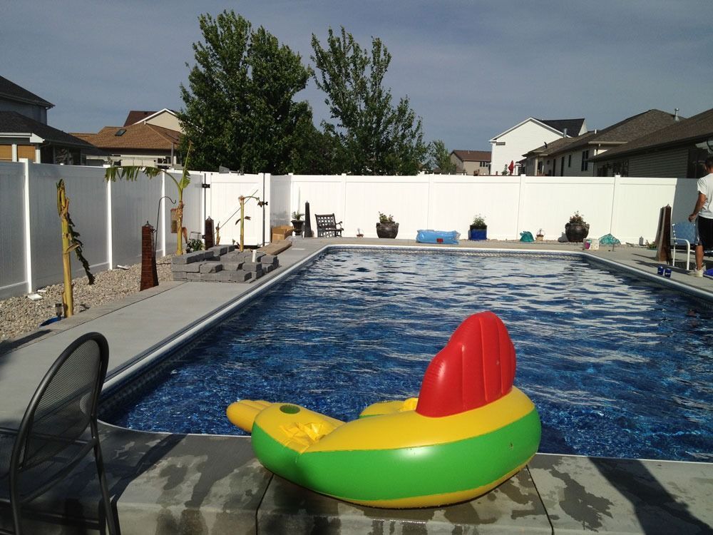 A swimming pool with an inflatable toy in front, surrounded by a white fence and houses on a sunny day.