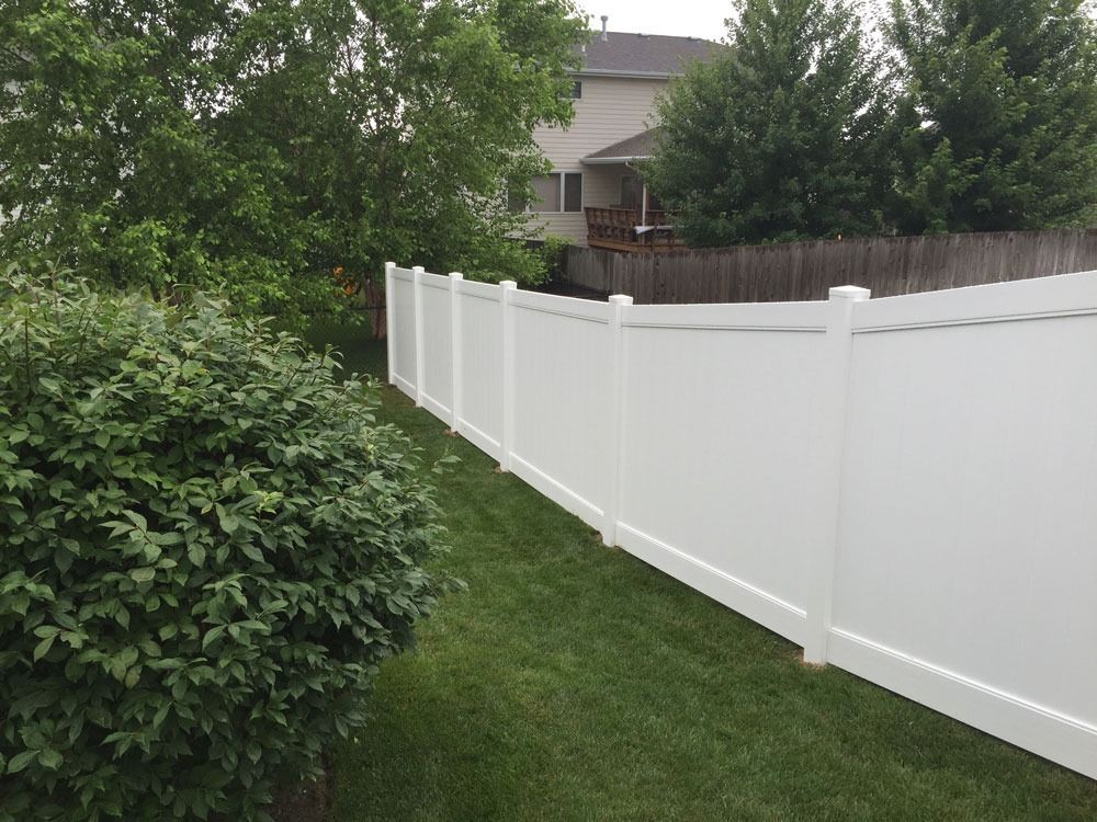 White vinyl fence in a grassy backyard, with a bush in the foreground and trees in the background.