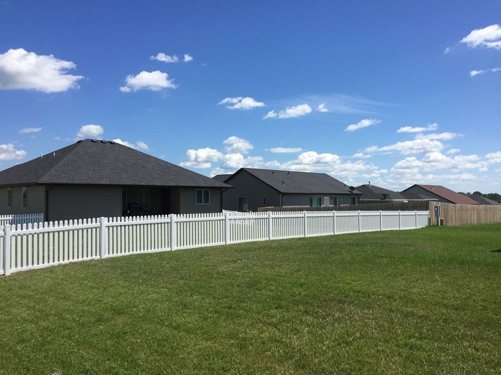 White picket fence in a grassy backyard, houses in background, blue sky with clouds.