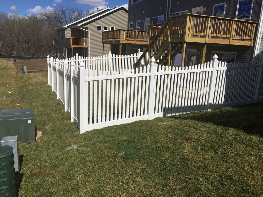 White picket fence surrounding a backyard with a grassy lawn; a house with a wooden deck is in the background.
