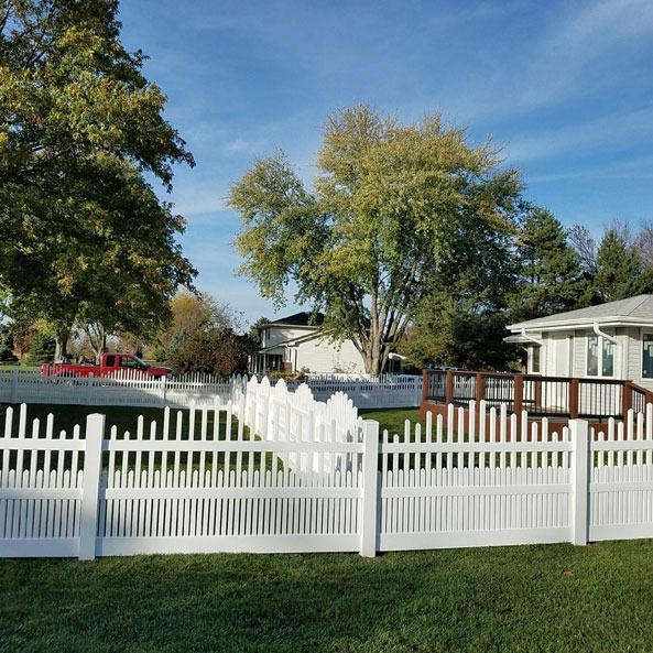 White picket fence in front of a house, sunny day, blue sky, green grass, and trees.