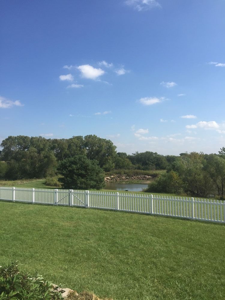 Green lawn, white picket fence, trees, and lake under a blue sky with clouds.