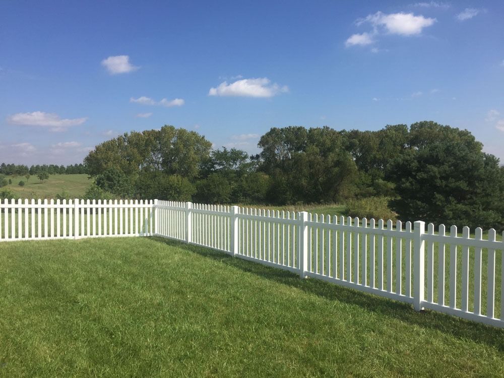 White picket fence on green lawn, rolling hills, and trees under a blue sky with clouds.