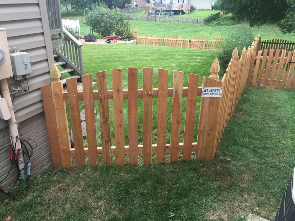 Wooden picket fence in a backyard; grass, electrical box on wall, gate.