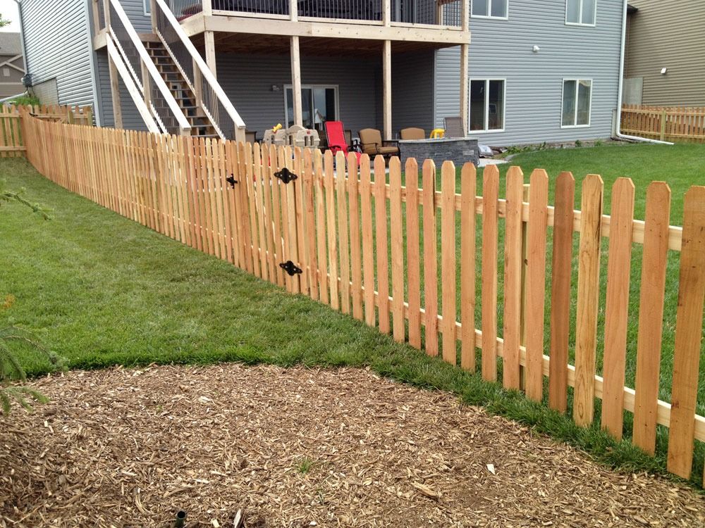 Wooden picket fence surrounds a backyard with a house in the background and green grass in the foreground.