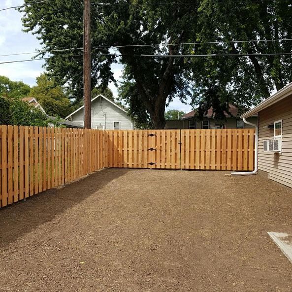 Wooden fence encloses a backyard with a gate, house on the right, and trees in the background.