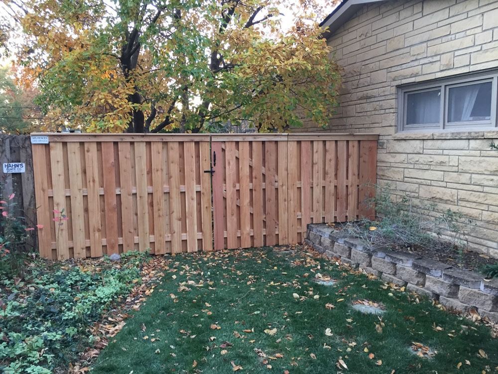 Wooden fence with gate in a yard. Green grass and fall foliage in background. Building on right.