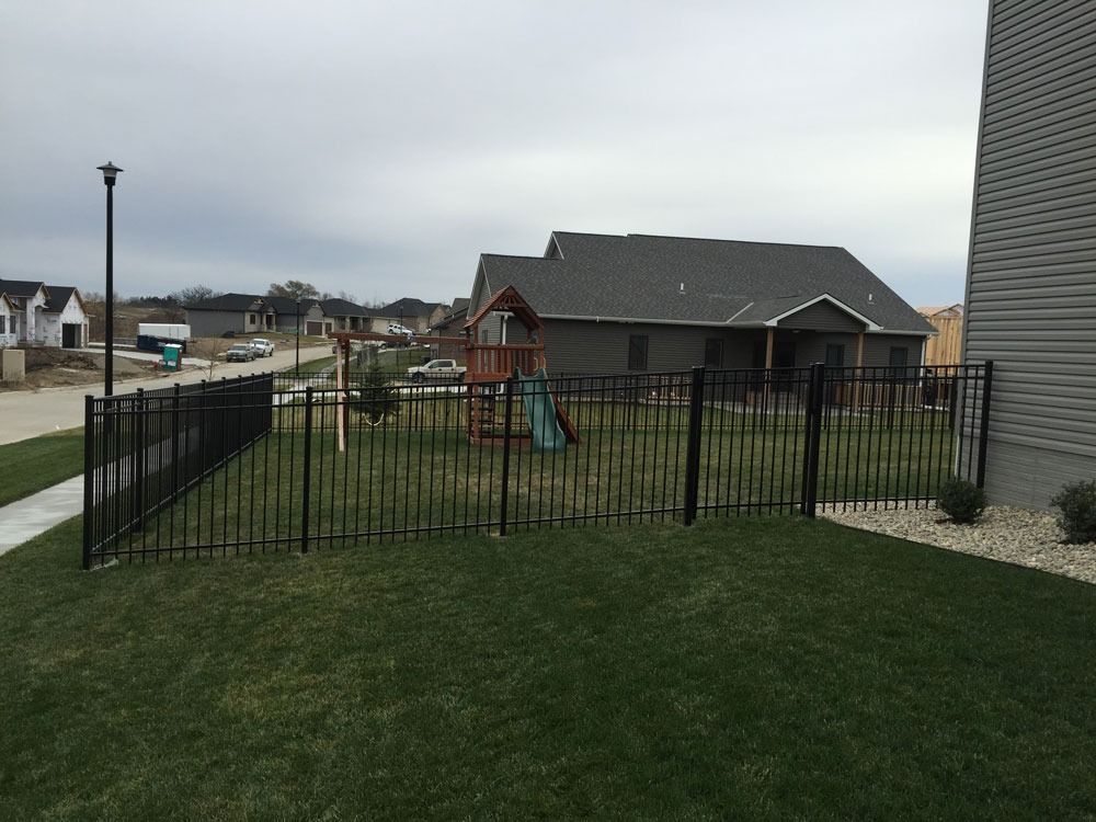 Black fence encloses a green lawn with a playground, backyard of a house on a cloudy day.