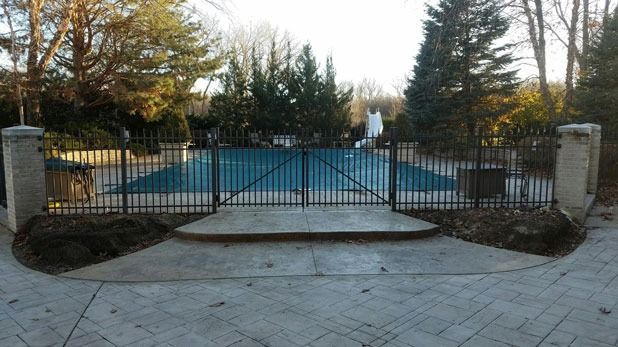 Black fenced pool entrance with concrete walkway, gate, and stone columns. Blue pool cover.