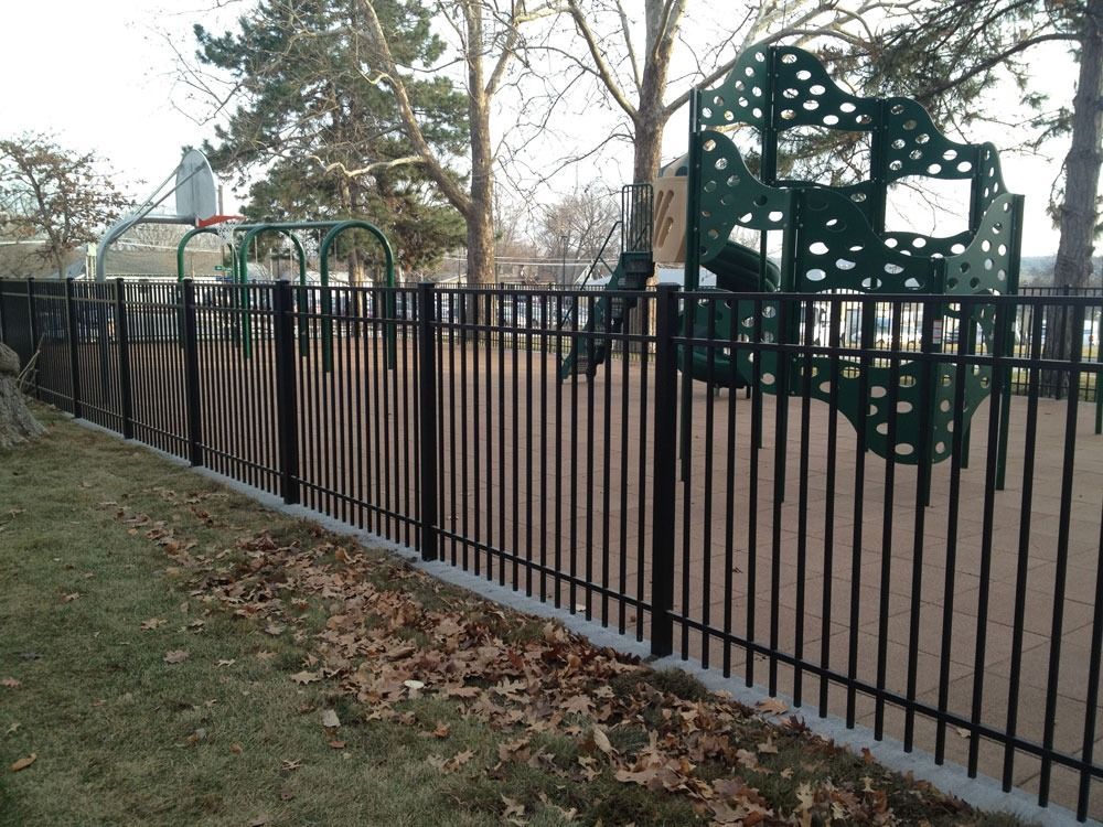 Black metal fence encloses a playground with a green climbing structure and basketball hoop.