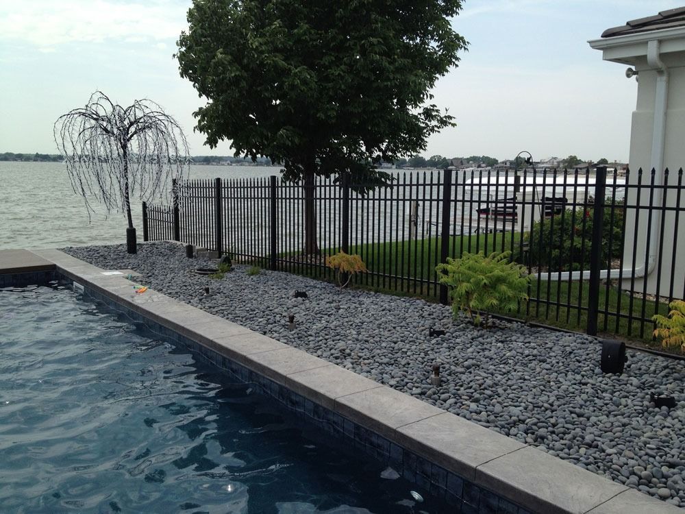 Poolside view of a lake behind a black fence, with a weeping willow-shaped sculpture and a tree.