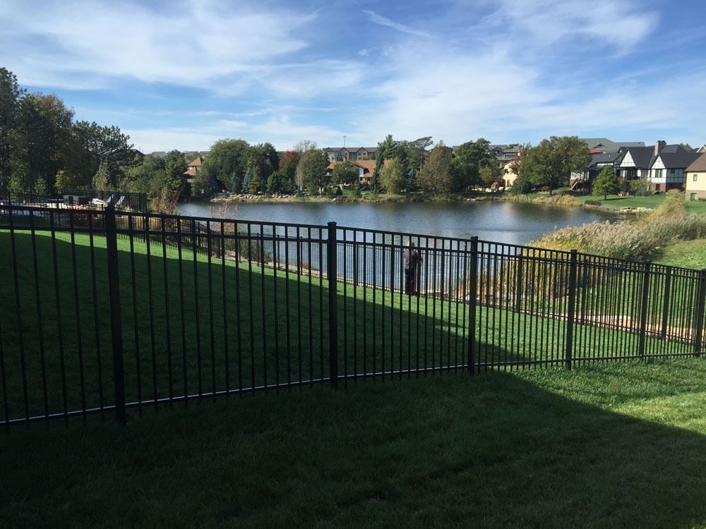 Black fence surrounds green grass, overlooking a lake with trees and houses under a partly cloudy sky.