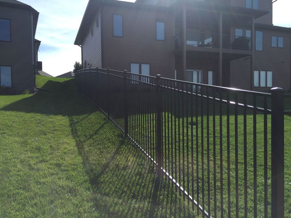 Black metal fence on a grassy hill, separating two brown houses. Sunny day, shadows cast.