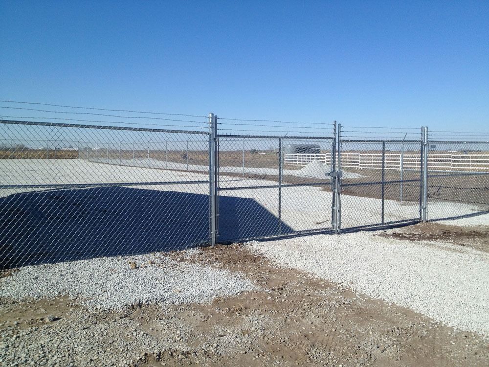 Chain-link fence and gate in a gravel area on a sunny day. Barbed wire extends above the fence.
