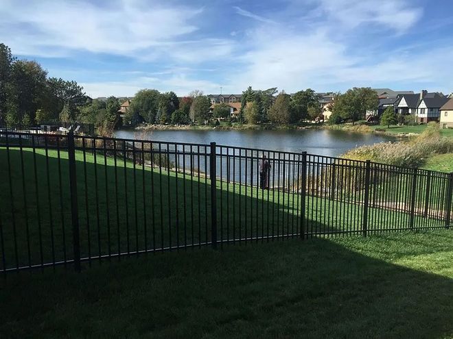 Black fence encloses green yard with a lake view, trees, and houses under a partly cloudy sky.