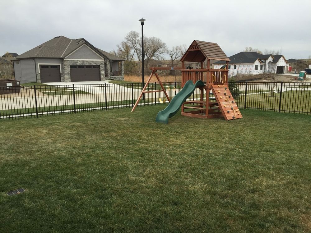 Backyard with a wooden playset, swings, and green grass. Houses and a black fence are in the background.