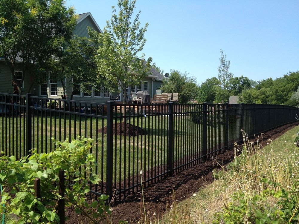 Black metal fence surrounding a backyard with grass, trees, and a house under a clear, blue sky.