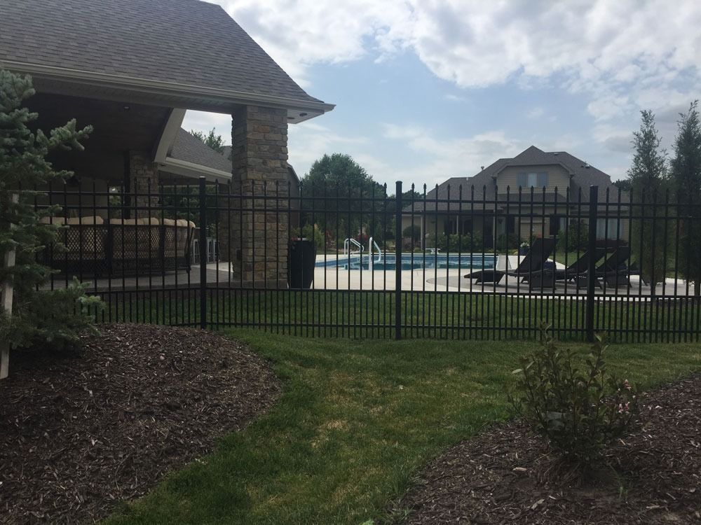 Black metal fence around a pool and patio. Stone pillar and large house in the background.