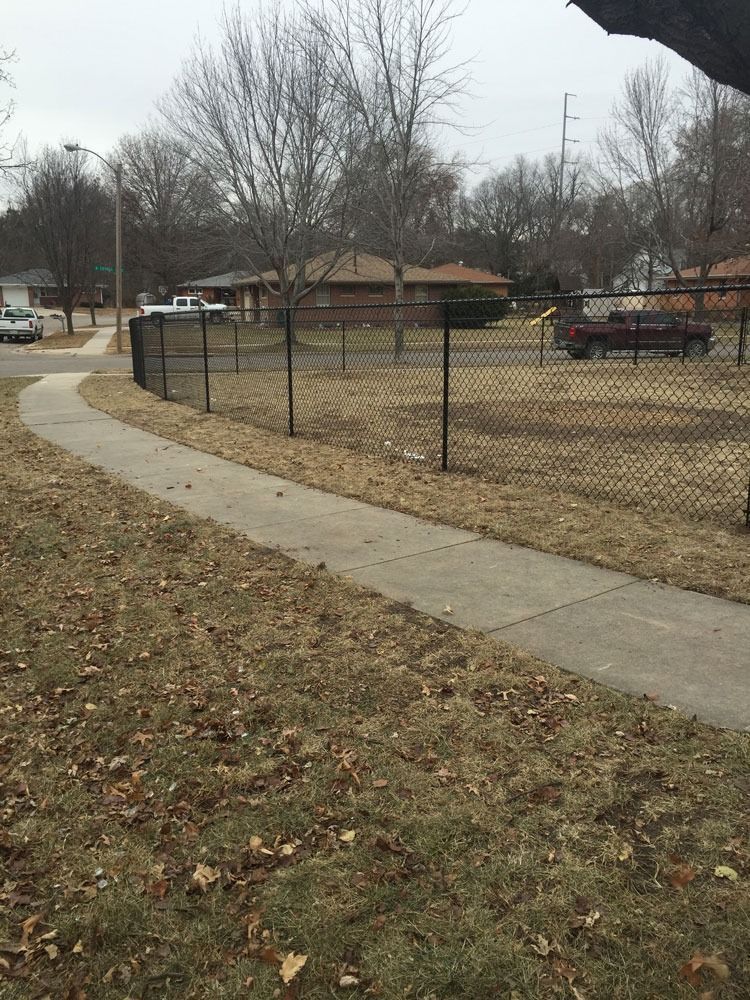 Sidewalk curves past a black chain-link fence, with trees and houses in the background on a cloudy day.