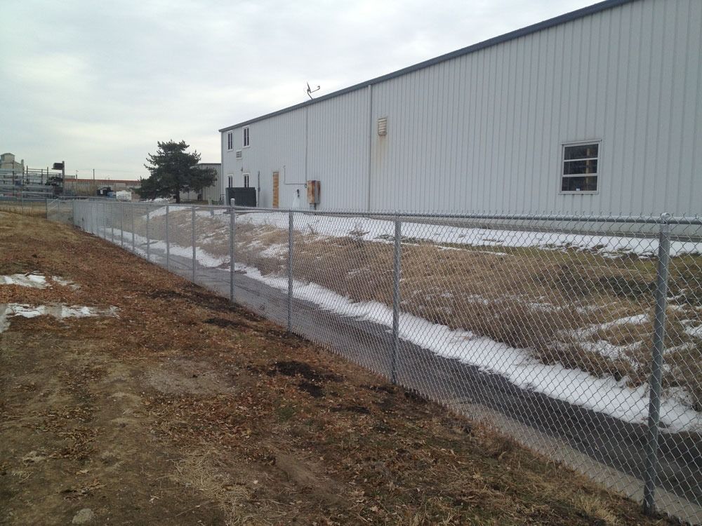 Chain-link fence runs alongside a building with white siding and a ditch with patches of snow on a cloudy day.