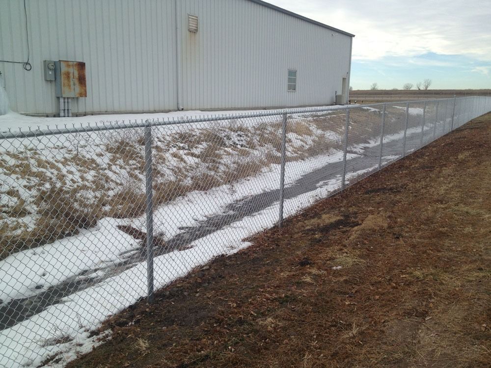 Chain-link fence borders a drainage ditch with snow, next to a white industrial building.