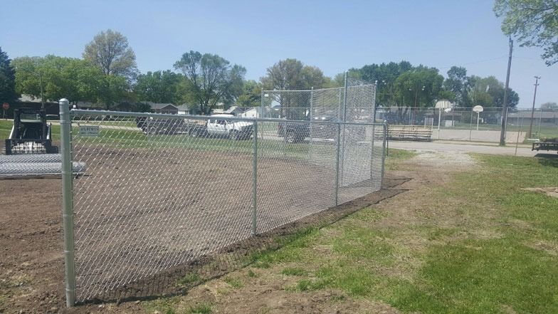 Chain-link fence surrounding a dirt area on a grassy lot, with construction vehicles and trees in the background.
