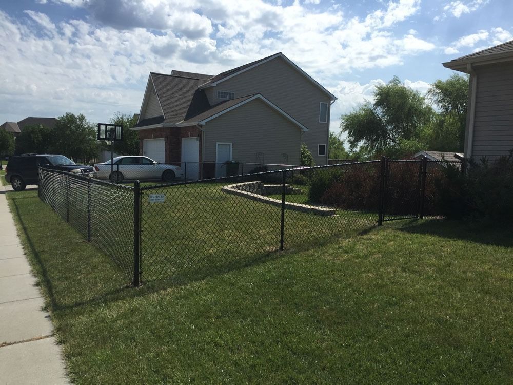 Black chain-link fence in front of a house. Cars and a garage are visible. Green grass and blue sky.