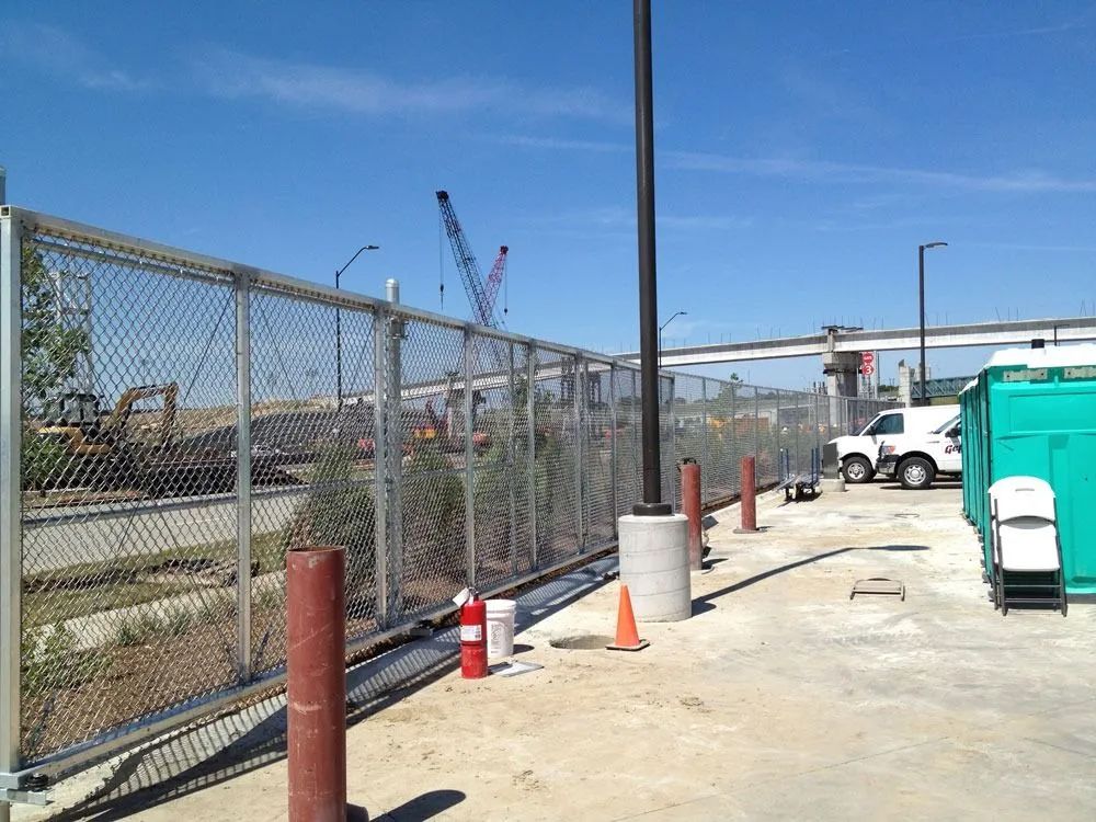 Chain-link fence encloses a construction site with equipment, trees, and portable toilets on a sunny day.